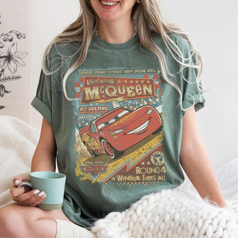 Pu&ograve; includere: T-shirt verde con grafica di Saetta McQueen del film Cars. Il design include il testo "Lightning McQueen" e "Round 4 Winner Takes All". Le maniche sono arrotolate.