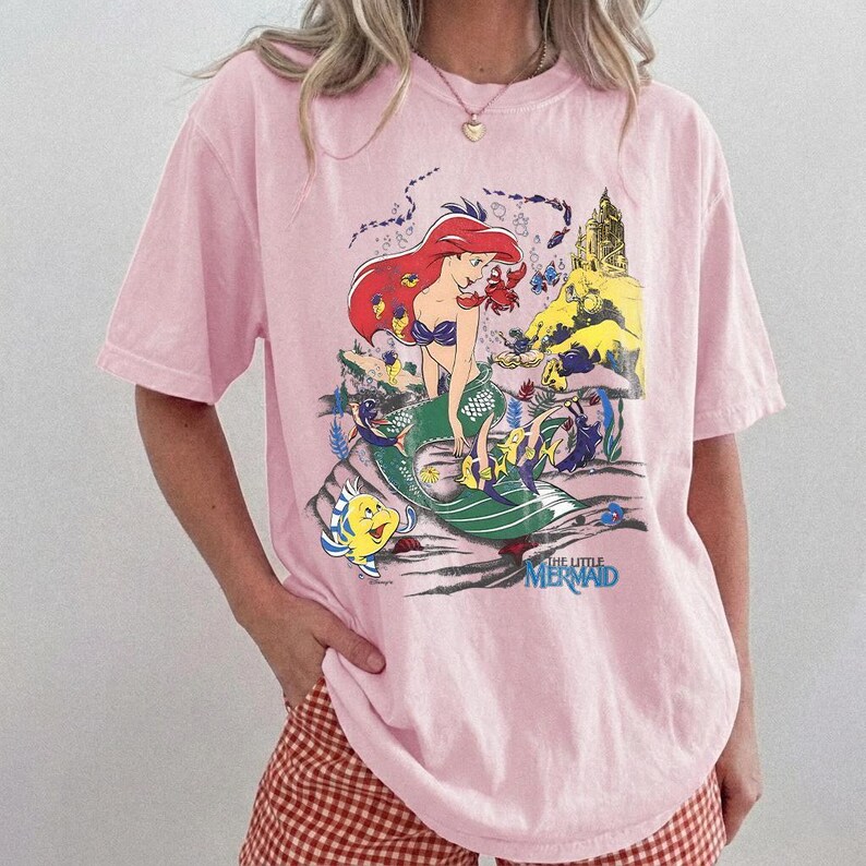May include: A light pink short-sleeve t-shirt featuring a graphic print of the character Ariel from The Little Mermaid. The design includes Ariel with red hair, a green tail, a yellow fish, a red crab, and a castle. The text The Little Mermaid is printed in blue at the bottom of the graphic.