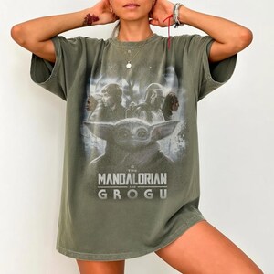 May include: Olive green t-shirt featuring a graphic of Grogu from The Mandalorian series. The design includes characters and the text "THE MANDALORIAN AND GROGU" in white. The shirt has short sleeves and a relaxed fit.