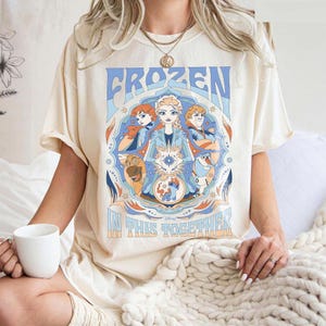 May include: Cream-colored t-shirt featuring a graphic from the Disney movie Frozen. The design includes the word "FROZEN" in blue, and the phrase "IN THIS TOGETHER". The graphic showcases characters Anna, Elsa, Kristoff, and Olaf.