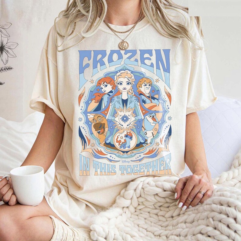May include: Cream-colored t-shirt featuring a graphic from the Disney movie Frozen. The design includes the word "FROZEN" in blue, and the phrase "IN THIS TOGETHER". The graphic showcases characters Anna, Elsa, Kristoff, and Olaf.
