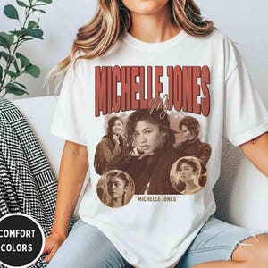 May include: White t-shirt featuring a sepia-toned image of a person with the text "MICHELLE JONES" in a red retro font. The shirt also has the text "Michelle Jones" in quotation marks.