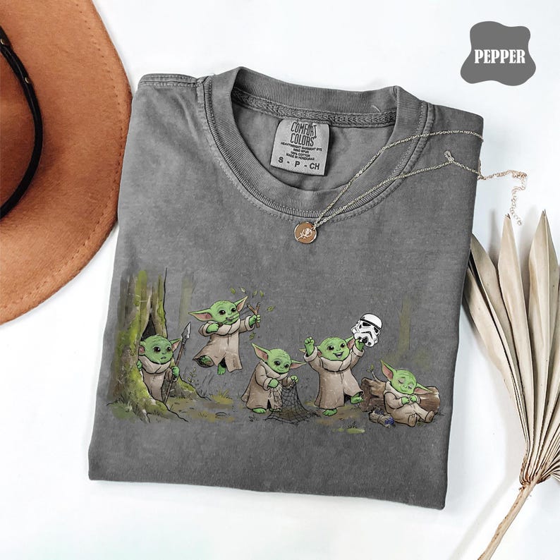 May include: A pepper-colored t-shirt featuring a graphic print of five small green creatures in robes. One holds a spear by a tree, one reaches up, one uses a net, one holds a white helmet, and one sits on a log. The shirt has a crew neck.