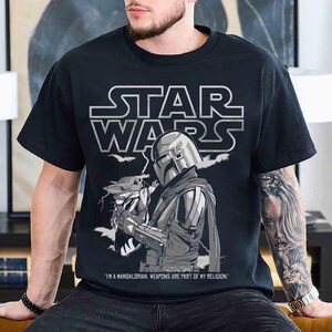 May include: Black Star Wars t-shirt featuring a graphic of The Mandalorian holding Grogu, with the Star Wars logo above. The text reads "I'm a Mandalorian. Weapons are part of my religion."