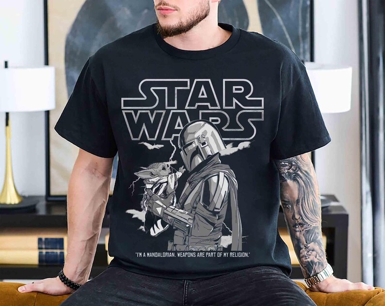 May include: Black Star Wars t-shirt featuring a graphic of The Mandalorian holding Grogu, with the Star Wars logo above. The text reads "I'm a Mandalorian. Weapons are part of my religion."