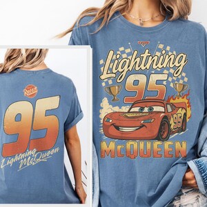 May include: A blue t-shirt featuring a graphic of the character Lightning McQueen with the text Lightning 95 McQueen and trophies. The back of the shirt displays the number 95 in a gradient orange font with the text Lightning McQueen below it.