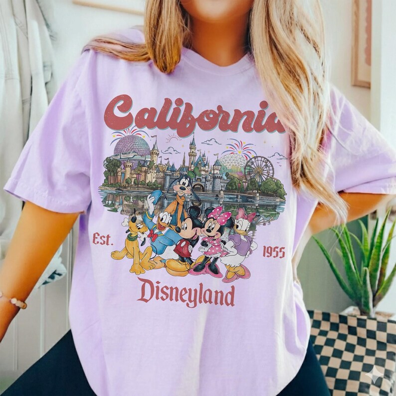 Może przedstawiać: Jasnofioletowa koszulka z grafiką Disneyland. Grafika zawiera napis "California" w czerwonym skrypcie, zamek Disneyland i postacie z kresk&oacute;wek. Tekst "Est. 1955" i "Disneyland" są r&oacute;wnież dołączone.