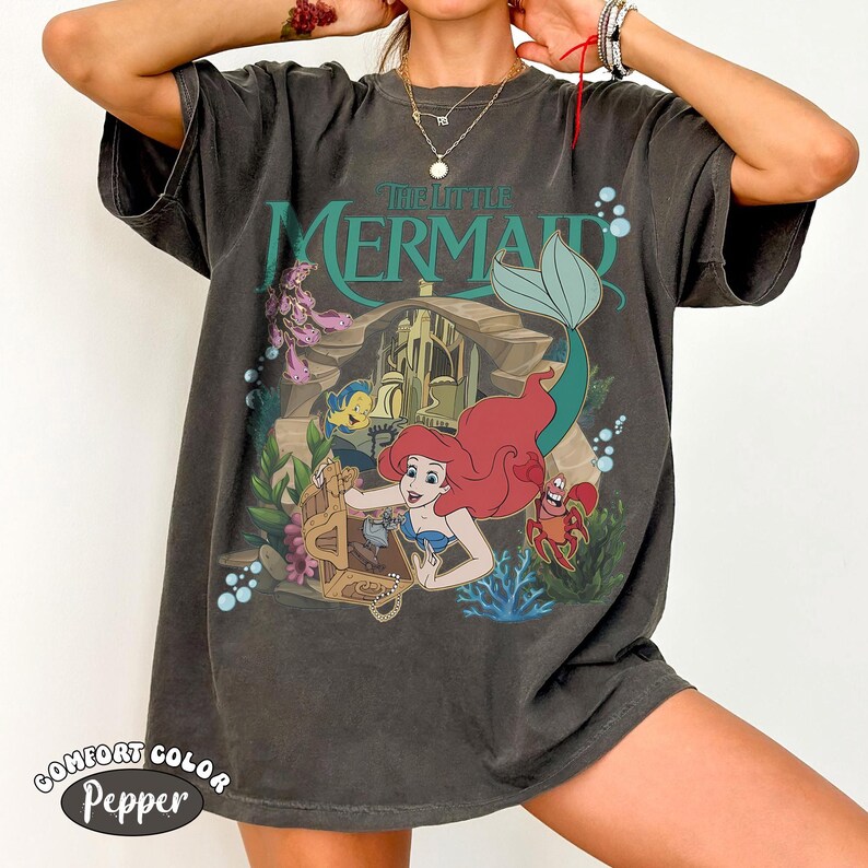 May include: A charcoal grey short-sleeve t-shirt featuring a graphic print of The Little Mermaid. The design displays Ariel, Flounder, and Sebastian near a treasure chest and underwater castle, with the text The Little Mermaid printed above.