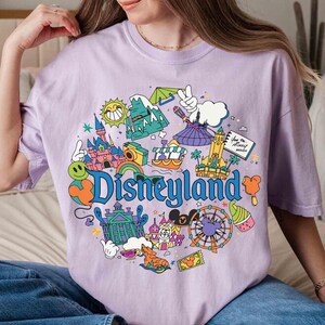 May include: A light purple t-shirt featuring a colorful Disneyland graphic. The design includes a castle, a Ferris wheel, and the word "Disneyland" in blue. The shirt is a comfortable, casual style.