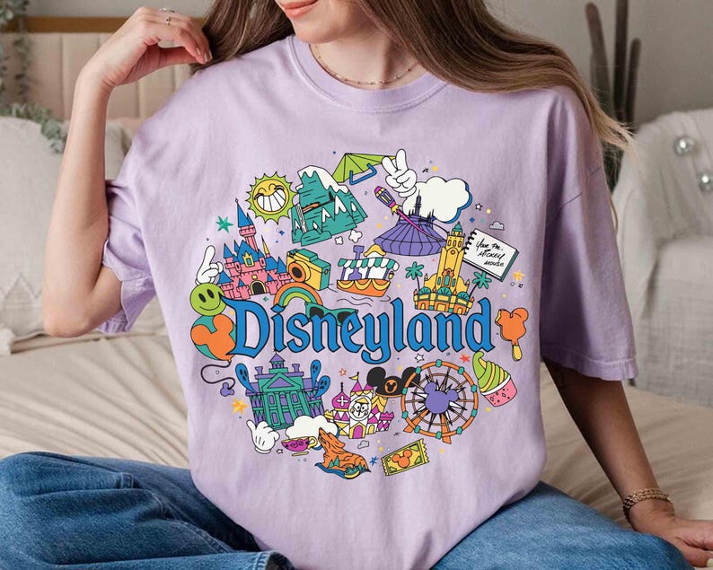 May include: A light purple t-shirt featuring a colorful Disneyland graphic. The design includes a castle, a Ferris wheel, and the word "Disneyland" in blue. The shirt is a comfortable, casual style.