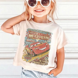 Pu&ograve; includere: T-shirt beige chiaro con una grafica di Saetta McQueen del film Cars. La grafica include il testo "Lightning McQueen" e "Round 4 Winner Takes All". La maglietta ha maniche corte.