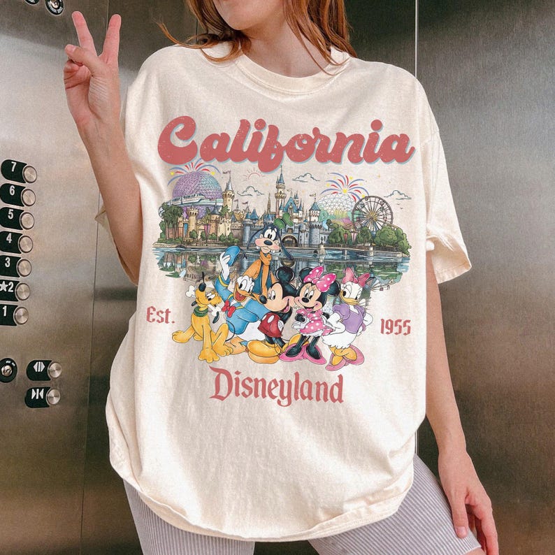Może przedstawiać: Kremowa koszulka z grafiką Disneylandu. Projekt zawiera słowo "California" w czerwonej, retro czcionce, zamek Disneylandu i r&oacute;żne postacie Disneya. Widoczny jest r&oacute;wnież tekst "Est. 1955" i "Disneyland".