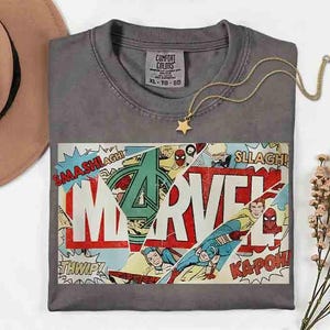 May include: A grey Comfort Colors t-shirt with a Marvel comic book design. The design features the Marvel logo, comic book action words, and superhero illustrations. A gold star pendant necklace is on the shirt.