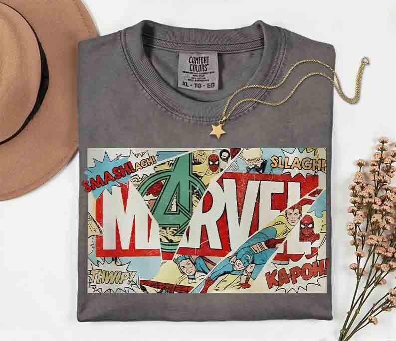 May include: A grey Comfort Colors t-shirt with a Marvel comic book design. The design features the Marvel logo, comic book action words, and superhero illustrations. A gold star pendant necklace is on the shirt.