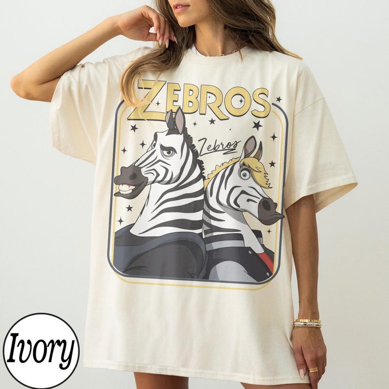 May include: Ivory-colored t-shirt featuring a cartoon graphic of two zebras with the word "ZEBROS" in yellow above them. The zebras are black and white with cartoonish features. The shirt has a relaxed fit.