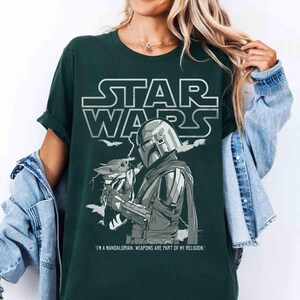 May include: Dark green t-shirt featuring a Star Wars graphic. The design includes the Star Wars logo, an illustration of The Mandalorian holding Grogu, and the quote "I'M A MANDALORIAN. WEAPONS ARE PART OF MY RELIGION."