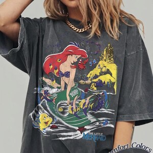 May include: A charcoal gray oversized t-shirt featuring a graphic print of Ariel from The Little Mermaid, surrounded by underwater characters like Flounder and Sebastian, with a castle in the background and the text The Little Mermaid and Comfort Colors Pepper.