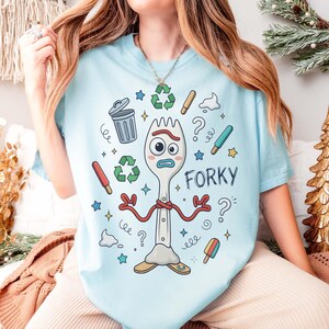 May include: Light blue t-shirt featuring a cartoon fork character named Forky. The design includes recycling symbols, question marks, and other playful elements. The word "Forky" is written in a playful font. A fun and whimsical graphic tee.