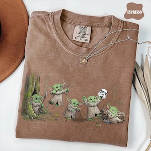 May include: An espresso-colored short-sleeve t-shirt featuring a graphic print of five small green creatures in a forest setting. One creature holds a spear, another a slingshot, one a net, one a stormtrooper helmet, and one is sleeping.