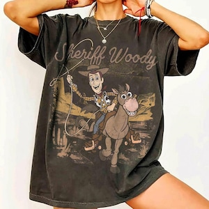 May include: Dark gray t-shirt featuring Sheriff Woody riding a horse, with the text "Sheriff Woody" in a vintage style. The graphic is in shades of brown, tan, and blue.