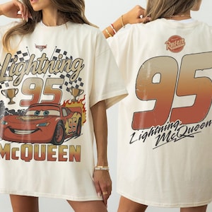 May include: An off-white t-shirt featuring a graphic of Lightning McQueen with the text Lightning 95 McQueen on the front. The back displays a large number 95 in a gradient orange and brown color with the text Rust-eze and Lightning McQueen.
