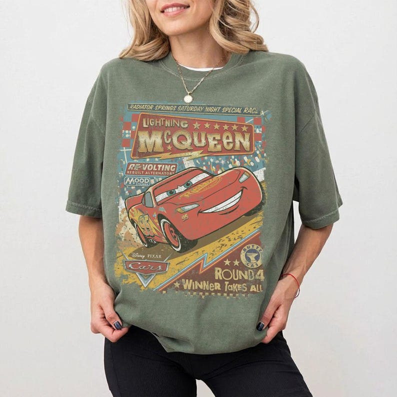 Pu&ograve; includere: T-shirt verde oliva con un motivo di Lightning McQueen, un'auto da corsa rossa, con la scritta "Lightning McQueen" e "Winner Takes All". Il design ha un aspetto vintage e usurato.