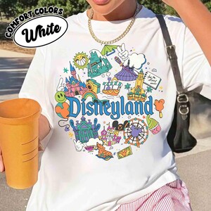May include: White t-shirt with a colorful Disneyland graphic. The design includes illustrations of a castle, a Ferris wheel, and other theme park icons. The words "Comfort Colors" and "White" are printed on the top left.