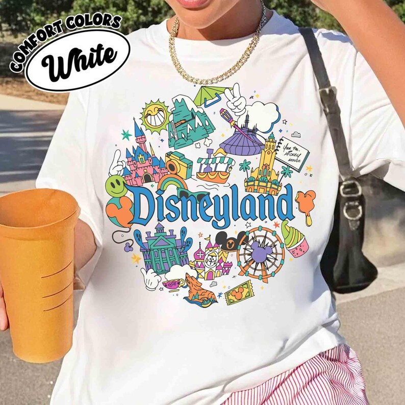 May include: White t-shirt with a colorful Disneyland graphic. The design includes illustrations of a castle, a Ferris wheel, and other theme park icons. The words "Comfort Colors" and "White" are printed on the top left.