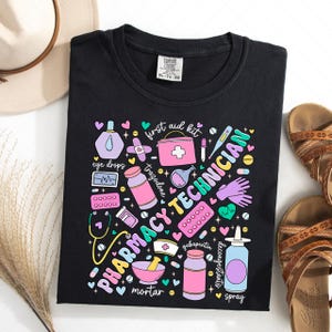 May include: Black t-shirt with a colourful graphic design featuring the words "Pharmacy Technician" surrounded by various pharmacy-related items, including a mortar and pestle, pills, eye drops, a first aid kit, and a stethoscope.