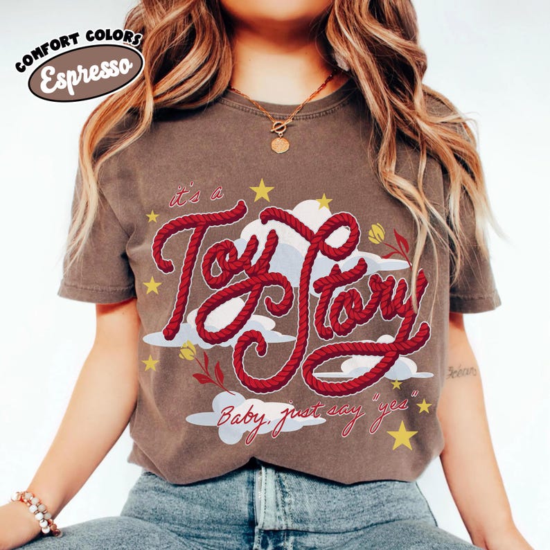 May include: A dark brown short-sleeve t-shirt featuring a graphic design with the red rope-style text "it's a Toy Story" and "Baby, just say yes". The design includes white clouds, yellow stars, and small red flowers on a dark brown background.