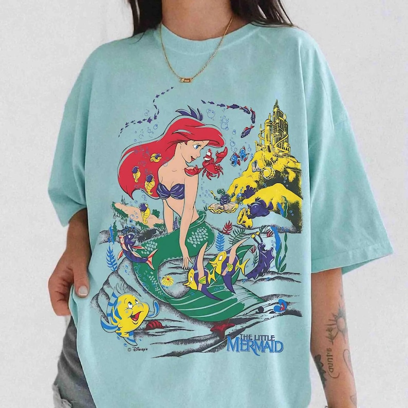 May include: A light blue t-shirt featuring a graphic print of Ariel from The Little Mermaid with red hair, a green tail, and various sea creatures including Flounder and Sebastian. The design includes a yellow castle and the text The Little Mermaid.