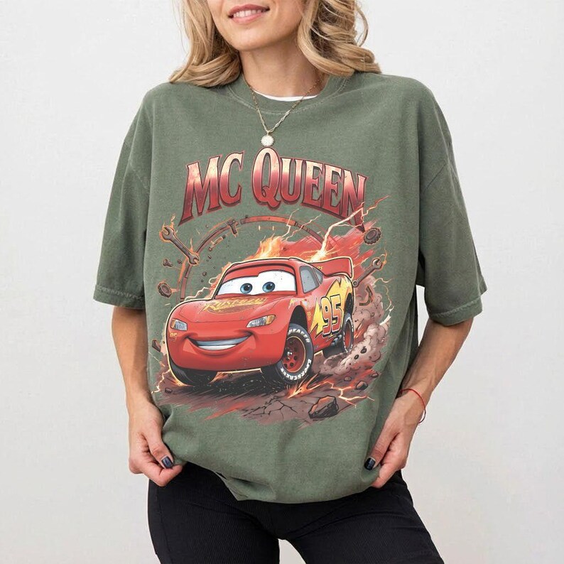 May include: An olive green t-shirt featuring a graphic of Lightning McQueen from the movie Cars. The design includes the text "MC QUEEN" in a red and orange gradient, with the number "95" on the car.