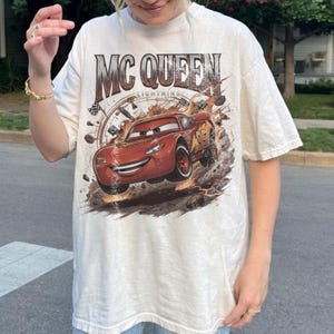 May include: An off-white short-sleeve t-shirt featuring a large graphic print of the red race car Lightning McQueen. The text MC QUEEN LIGHTNING is printed above the car, which is surrounded by racing-themed elements like tyres and flags.