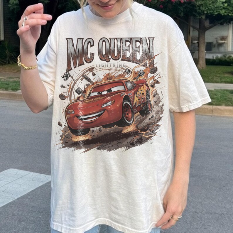 May include: An off-white short-sleeve t-shirt featuring a large graphic print of the red race car Lightning McQueen. The text MC QUEEN LIGHTNING is printed above the car, which is surrounded by racing-themed elements like tyres and flags.