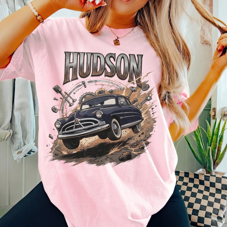 May include: A light pink t-shirt featuring a graphic print of a dark blue vintage car with eyes on the windscreen. The word HUDSON is printed in large, bold, metallic-toned letters above the car, surrounded by mechanical gears and engine parts.
