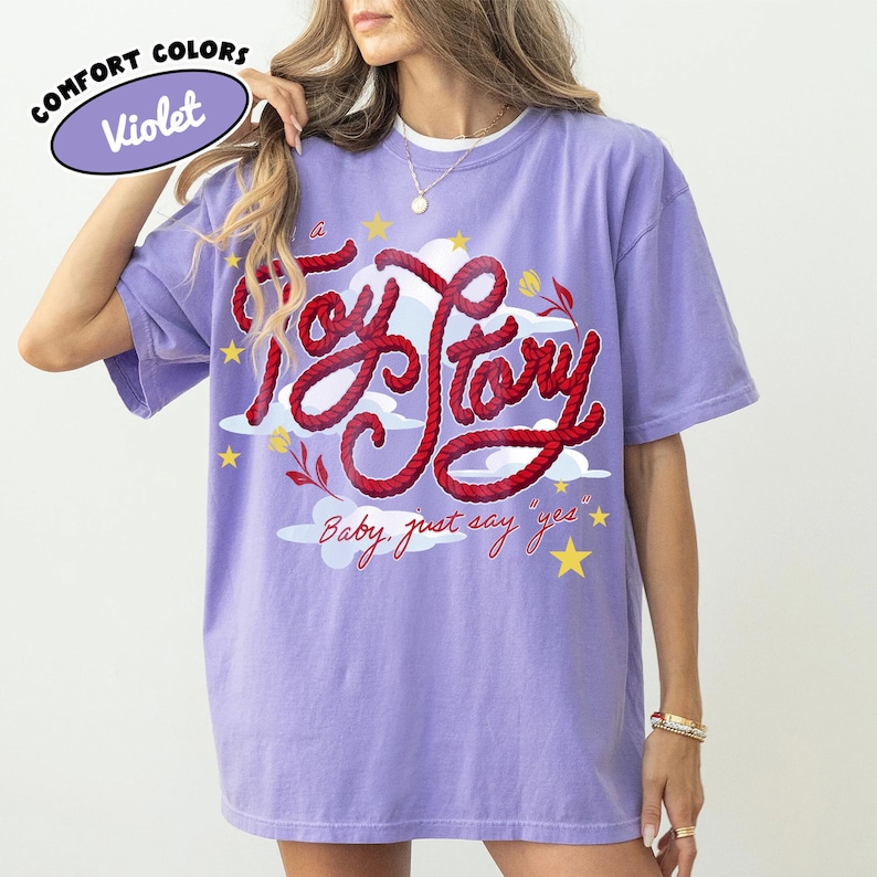 May include: A violet short-sleeve t-shirt featuring a graphic print of the red rope-style text Toy Story, yellow stars, white clouds, and small red flowers. Below the main text, the phrase Baby, just say yes is printed in red cursive script.