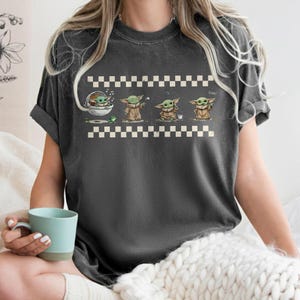 May include: Dark gray t-shirt featuring a cartoon illustration of a character in various poses. The design includes a series of images of the character with a checkered border and the word "Cool" printed on the shirt.