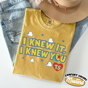 Puede incluir: Una camiseta de manga corta color amarillo mostaza con el texto azul I KNEW IT, I KNEW YOU, un coraz&oacute;n rojo con las letras TS y un gr&aacute;fico de pelota de colores. La camiseta incluye nubes blancas y estrellas azules, con etiqueta Comfort Colors.