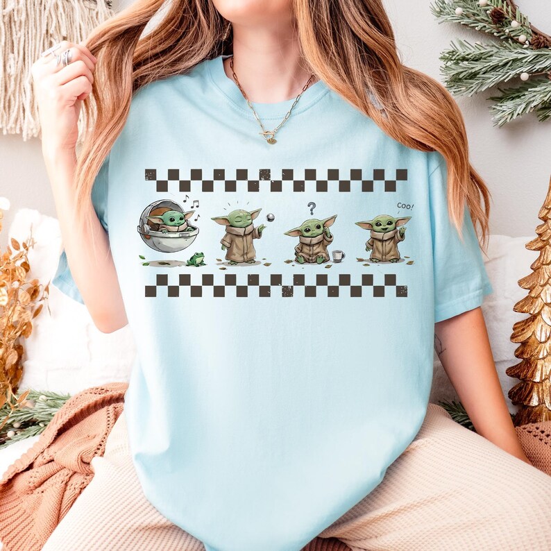 May include: Light blue t-shirt featuring a graphic of a character from a popular science fiction series. The graphic shows the character in four different poses, with the word "Cool" printed on the right side. The shirt has a checkered pattern at the top and bottom.