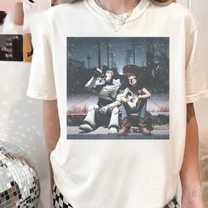 May include: Cream-colored t-shirt featuring a distressed graphic of Buzz Lightyear and Woody from Toy Story. The characters are depicted sitting on a curb, with a vintage, faded aesthetic. The shirt has a classic crew neck and short sleeves.