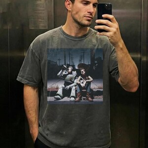 May include: A gray t-shirt featuring a graphic of Buzz Lightyear and Woody from Toy Story. The image shows the characters sitting together in a street scene. The t-shirt is a relaxed fit.