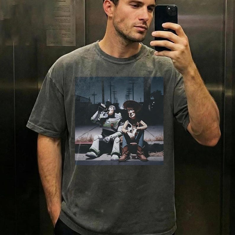 May include: A gray t-shirt featuring a graphic of Buzz Lightyear and Woody from Toy Story. The image shows the characters sitting together in a street scene. The t-shirt is a relaxed fit.