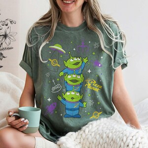 May include: Green t-shirt featuring three green aliens in blue suits, with the text "Pizza Planet". The shirt also has graphics of a spaceship, a rocket, and other space-themed elements.