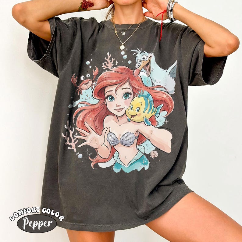 Op de afbeelding: Een antracietgrijs t-shirt met korte mouwen en een grafische print van een zeemeermin met rood haar, een gele en blauwe vis, een kleine krab en een witte meeuw. Het ontwerp bevat koraal- en bubbelaccenten. De tekst Comfort Color Pepper is zichtbaar.