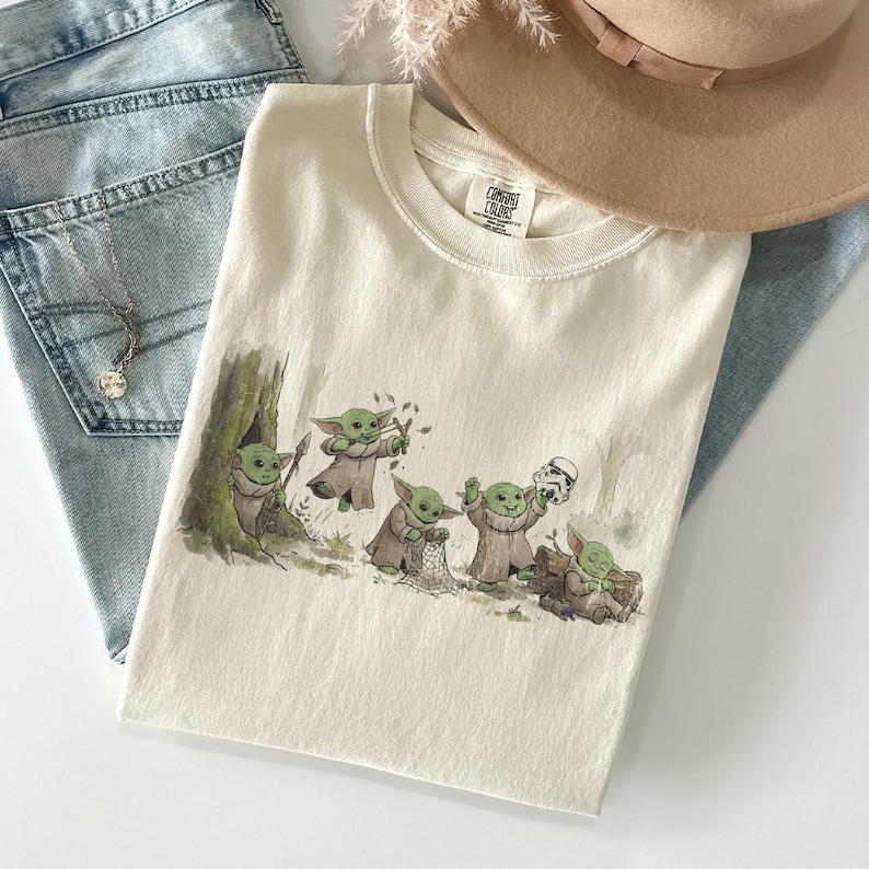 May include: A cream-colored t-shirt featuring a graphic illustration of five small green creatures in robes. One creature peeks from a tree, one uses a slingshot, one holds a net, one holds a white helmet, and one sits on a log. The shirt has a Comfort Colors label.