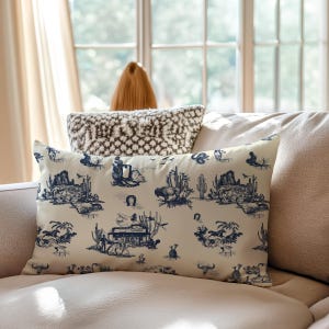 Western toile lumbar pillow in cream and navy blue styled on a beige sofa in soft daylight, featuring cowboy, buffalo, cactus, and saloon motifs