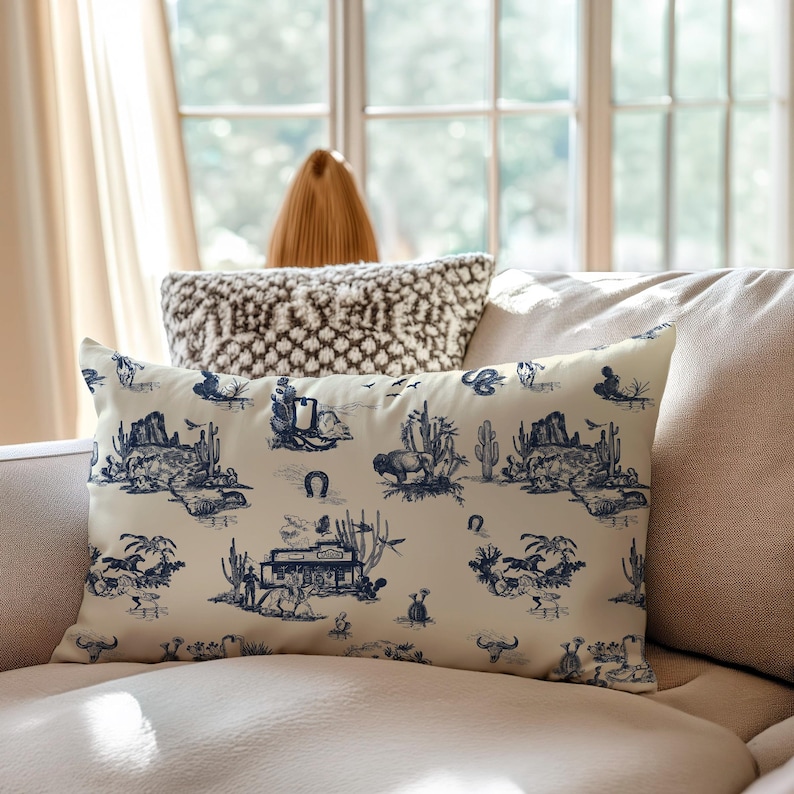 Western toile lumbar pillow in cream and navy blue styled on a beige sofa in soft daylight, featuring cowboy, buffalo, cactus, and saloon motifs