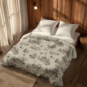 May include: A bed featuring a white duvet cover with a repeating dark brown toile-style print depicting desert scenes, including cacti, horseshoes, and small buildings. Two plain white square pillows are placed at the head of the bed.