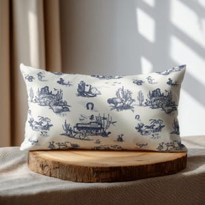 Western toile lumbar pillow in cream and navy blue displayed on a round wooden pedestal with soft natural window shadows