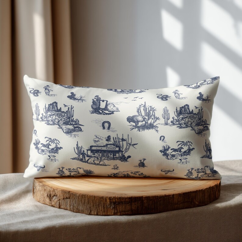 Western toile lumbar pillow in cream and navy blue displayed on a round wooden pedestal with soft natural window shadows